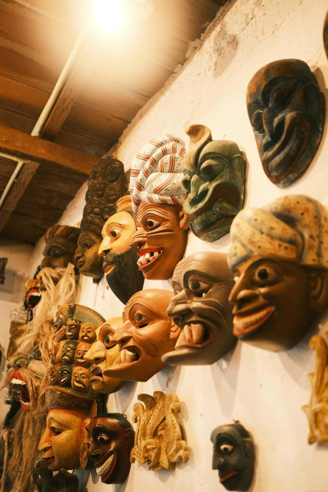 Carved and painted wooden masks hung in rows on a workshop wall under a bare bulb